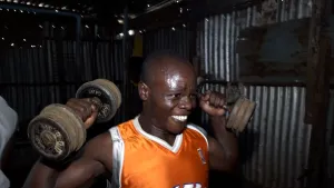 Een foto van een man in Kenia. Hij staat binnen, in een donkere ruimte, en tilt gewichten op. Hij is bezweet en kijkt ingespannen.