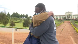 Een foto van de achttienjarige Brian uit Kenia. Hij heeft een mondmasker op en omhelst zijn moeder. Op de achtergrond zie je een groot gebouw.