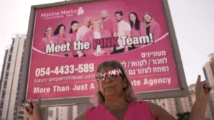 Een foto van pink lady Maxine. Zij is een makelaar in Israël. Ze heeft roze kleren aan en staat voor een groot reclamebord. Daar staat haar bedrijf op vermeld. Het bord is ook roze van kleur. Daarachter zie je flatgebouwen.