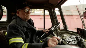 Een foto van een oude man in een oude brandweerwagen. Je ziet hem achter het stuur zitten en je ziet de binnenkant van de wagen. De man heeft een uniform aan.