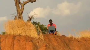 Een foto van een man die op een rots zit, met zijn laptop op schoot. De rots is roodbruin van kleur en erop groeit geel gras en een boom. De man heeft geen schoenen aan.