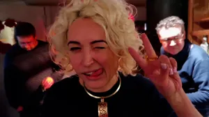 Een foto van een vrouw met blond haar. Ze kijkt in de camera en maakt het V-teken met haar hand. Ze steekt haar tong ietsje uit, naar de zijkant van haar mond. Op de achtergrond zie je twee mannen.