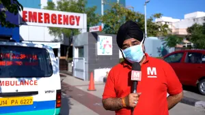 Een foto van een presentator die met een mondmasker op voor een ziekenhuis staat met een microfoon in zijn hand. Links is een ambulance te zien.