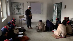 Een foto van meester Sharif in Pakistan. Hij staat in een klaslokaal. Op de grond zitten ongeveer tien mensen. De meester draagt een mondmasker. 