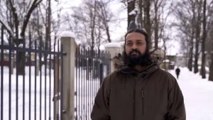 Een foto van een Indiase man die buiten staat. Hij heeft een zwarte baard, en draagt een winterjas. Op de grond ligt sneeuw. Naast hem zie je een hek, met daarachter bomen. 