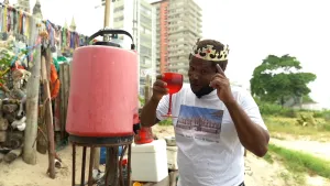 Een foto van een man die buiten staat. Hij heeft een kroon op zijn hoofd en een rood wijnglas in zijn hand. Hij kijkt in de camera. Naast hem staat een rood vat. Op de achtergrond zie je een flatgebouw.