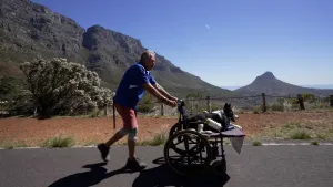 Een foto van een man die, op een weg, achter een rolstoel loopt. Op de rolstoel ligt zijn hond. Op de achtergrond zie je bergen, bomen en een blauwe lucht.