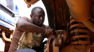 Een foto van een automonteur in Afrika. Hij is een auto aan het aanpassen. Je ziet een van de veren van een auto. Die zit bij de wielen.