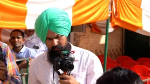Een foto van de Indiase influencer Darshan. Hij heeft een tulband op zijn hoofd. Dat is een Indiase hoed. Met zijn camera is hij iets aan het filmen. Hij kijkt aandacht naar het schermpje van zijn camera. Op de achtergrond zie je een oranje doek hangen.