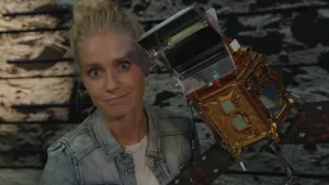 Een foto van presentator Janouk die naast een kleine versie staat van een ruimte satelliet.