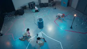 Een foto van vier kinderen in een ruimte met lichtblauw licht. Ze zitten ieder bij een met tape afgeplakt gedeelte op de vloer. Ze hebben allemaal een mandje met witte tafeltennisballen bij zich staan en die ballen liggen ook door de hele ruimte heen.