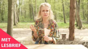 Een foto van Janouk die aan een tafel van lichtgekleurd hout zit in een bos. Ze heeft in haar handen een witte puntzak met rode vierkantjes erop. Het lijkt of ze een frietje of snack eet en erop kauwt met gesloten lippen. Aan haar vingers zit fritessaus. 