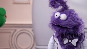 Een foto van een paarse pop die naar een groene pop kijkt waarvan links alleen het gezicht te zien is. 