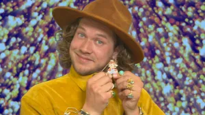 Een foto van presentator Pascal met een hoed op. Hij laat met zijn handen een halsketting zien van Super Mario die hij om zijn nek heeft. Hij heeft veel mooie ringen om zijn vingers.