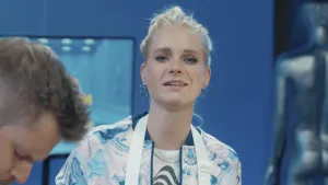 Een foto van presentator Janouk die in de studio staat en naar de camera kijkt. Links is het hoofd van presentator Job te zien die met iets bezig is.
