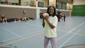 Een foto van presentator Tirsa die in een gymzaal staat met een bal in haar handen.