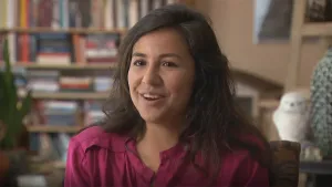 Een foto van een vrouw die in een kamer voor de camera zit. Op de achtergrond is er een boekenkast vol met boeken zichtbaar.