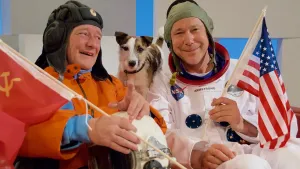 Een foto van acteurs die de astronauten Neill Armstrong en de Rus Yuri Gagarin spelen. Ze hebben beide astronauten pakken aan maar dragen geen helm. Er staat een hondje tussen hun in.