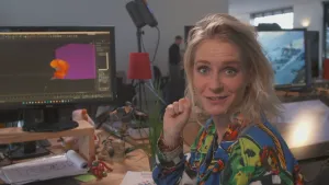 Een foto van presentator Janouk. Ze zit achter een bureau en kijkt in de camera. Achter haar zie je een computerbeeldscherm met daarop een oranje vorm. Op het bureau staan nog wat poppetjes. Dat zijn actiefiguren.