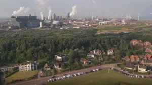 Een foto die vanaf grote hoogte gemaakt is. Op de voorgrond zie je huizen en geparkeerde auto's. Daarachter zie je een bos en daarachter een aantal grote fabrieken. Uit de schoorstenen daarvan komt veel witte rook.