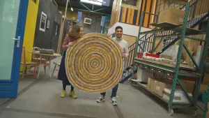 Een foto van presentator Nizar. Hij staat in een soort fabriekshal samen met kunstenaar Simone Post. Samen houden ze een rond kleed in de lucht. Het kleed heeft de kleuren geel en grijs.
