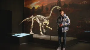 Een foto van presentator Nizar. Hij staat in een museum naast het skelet van een dinosaurus. Nizar kijkt in de camera. De dinosaurus staat op twee poten en heeft twee armen en een vrij lange nek.