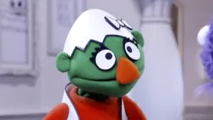 Een foto van een groene pop met een eierschaal op haar hoofd. Ze heeft een oranje neus en oranje kleren aan.