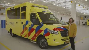 Een foto van presentator Pascal naast een grote gele ambulanceauto. Pascal heeft zijn hand op de motorkamp van de ambulance en vertelt iets aan de kijker. De ambulance staat in een grote garage voor ambulances.