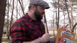 Een foto van boswachter Tim, in een bos. Hij heeft een bord van de Beestenbrigade bingo in zijn handen, en een hamer. 
