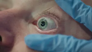 Een foto van een wijd opengesperd groen met blauw oog van een mens van dichtbij genomen. Het oog wordt opengesperd door twee vingers van een blauw gehandschoende hand. Het is het oog van prof en de hand van Janouk.