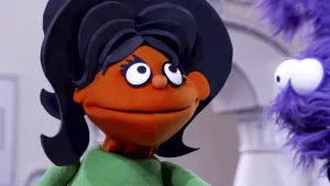 Een foto van een oranje pop met een grote bos zwart haar. Ze kijkt naar de presentator, dat is een blauwe pop.