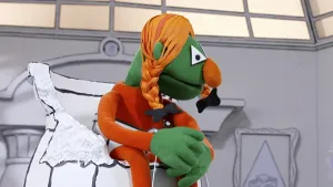 Een foto van een groene pop, met oranje kleren, en met oranje haar met vlechtjes. Ze zit op een stoel en kijkt opzij.