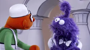 Een foto van een blauwe pop. Dat is de presentator. Tegenover hem staat een oranje pop met een witte hoed op. Ze kijken elkaar aan.