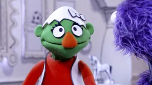 Een foto van een groene pop met een eierschaal op zijn hoofd. Op de voorgrond zie je nog een stukje van een blauwe pop. Dat is de presentator. 