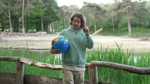 Een foto van presentator Pascal die een wereldbol vasthoudt. Hij staat in een dierentuin en legt iets uit aan de kijker. In de achtergrond zie je zebra's, bomen en water.