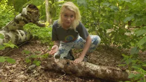 Een foto van presentator Janouk. Ze zit in een bos met een knie op de grond. Voor haar ligt een boomstam met zwammen erop. Ze legt wat uit naar de kijker. 
