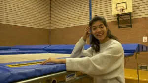 Een foto van een vrouw naast een trampoline. Ze staat in een sporthal en kijkt in de camera. Op de achtergrond zie je een basket van basketbal.