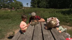 Een foto van boswachter Tim die op een houten bank zit, samen met een jongetje en een meisje. Op tafel staat een glazen bak met water erin. Ze kijken alle drie naar de diertjes in die bak. Op de achtergrond zie je een meertje, met bomen. 