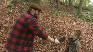 Een foto van boswachter Tim in het bos. Op de grond liggen veel herfstbladeren. Voor Tim staat een jongetje. Tim houdt een stuk hout voor het gezicht van het jongetje alsof het een microfoon is. 