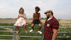 Een foto van boswachter Tim. Hij staat in de natuur, voor een ijzeren hek. Op dat hek zitten twee jonge meisjes. Tim is iets aan het vertellen. Op de achtergrond zie je weilanden en bomen.