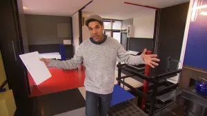 Een foto van de presentator in een grote open ruimte in een huis. De vloer en muren hebben grote vlakken in zwart, wit, grijs, rood en blauw. De presentator staat met zijn armen wijd te praten in de camera.