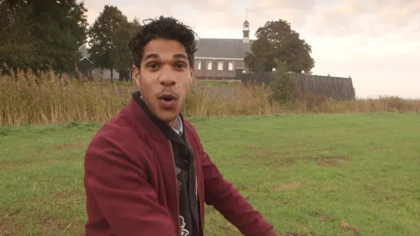 Een foto van de presentator die in een weiland staat. Achter hem is een verhoogd stuk grond met daarop een kerk. Naast de kerk staan bomen.