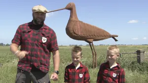 Een foto van boswachter Tim. Hij zit staat in een weiland. Naast hem staan twee jongetjes. Tim praat in de camera. Achter hen staat een metershoog houten beeld van een grutto. Dat is een vogel.