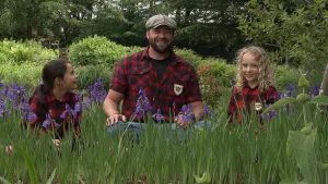 Een foto van boswachter Tim. Hij zit buiten op de grond. Links en rechts van hem zit een meisje. Ze glimlachen alle drie. Op de voorgrond zie je paarse bloemen. Op de achtergrond zie je allemaal planten en bomen.