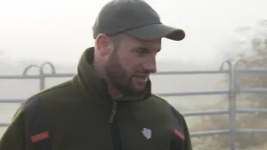 Een foto van een boswachter met een pet op. Hij staat voor een ijzeren hek en kijkt opzij. In de verte zie je mist.