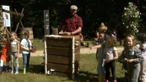 Een foto van boswachter Tim. Hij staat buiten achter een houten tafel. Links en rechts van hem staan kinderen. Op de achtergrond zie je links in beeld een bord met de foto van een bever. De zon schijnt.