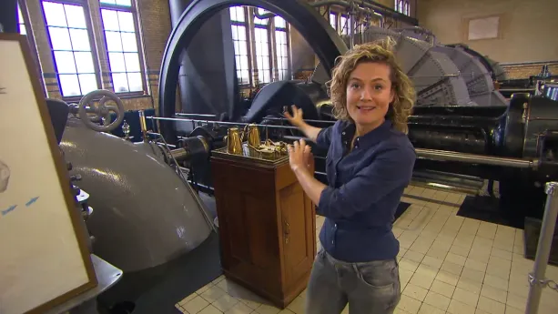 Een foto van de presentator Nienke in een ruimte met grote machines en buizen. Ze wijst naar achteren, naar een groot wiel dat met stangen en buizen vastzit aan de rest. Achter het wiel staan even grote wielvormige kasten.