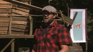 Een foto van boswachter Tim. Hij staat buiten en vertelt iets. Hij heeft een rood-zwart geruit overhemd aan en een pet op. Achter hem zie je een bord met daarop de foto van een vos. Daarnaast zie je een boomhut.