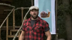 Een foto van boswachter Tim. Hij staat buiten en praat tegen de camera. Hij heeft een rood-zwart geruit overhemd aan en een pet op. Achter hem zie je een bord met daarop de foto van een slak.