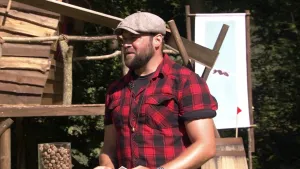 Een foto van boswachter Tim. Hij staat buiten en kijkt opzij. Hij heeft een rood-zwart geruit overhemd aan en een pet op. Op de achtergrond zie je een boomhut en een bord met daarop de foto van een mier.